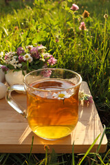 Cup of aromatic herbal tea, pestle and ceramic mortar with different wildflowers on wooden board in meadow
