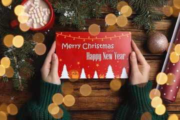 Woman holding greeting card at wooden table, top view. Christmas celebration