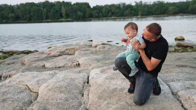 Father holding his little baby boy on one knee. Loving daddy kissing his son's cheek and back of the neck. Family concept. River at backdrop.
