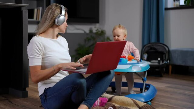 Positive busy Caucasian woman in headphones talking using video chat on laptop as curios infant in baby walker walking at background. Confident mother messaging online working as kid enjoying leisure