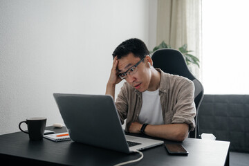 Middle aged surprised asian man working at home with laptop.