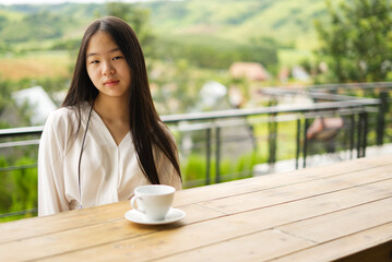 An Asian girl with a cup of coffee on a wooden table and copy space.  An Asian girl on the balcony with green trees and mountains in the background from Thailand.
