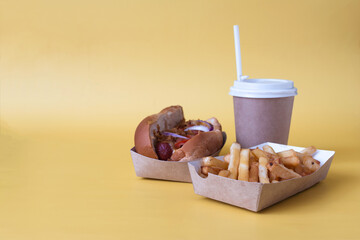 A hot dog with sausage, French fries and a drink on a yellow copy space background.