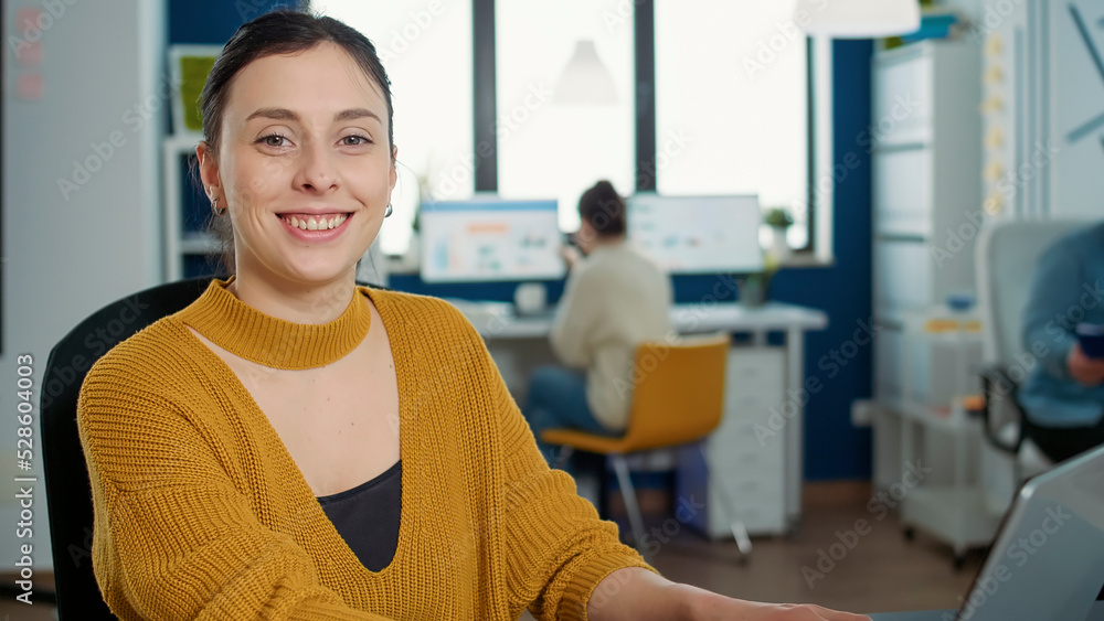 Poster portrait of woman typing using laptop keyboard and smiling at camera sitting at desk in busy startup
