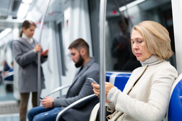 Obraz premium Adult woman browsing and typing messages on tablet in subway car leaning on handrail