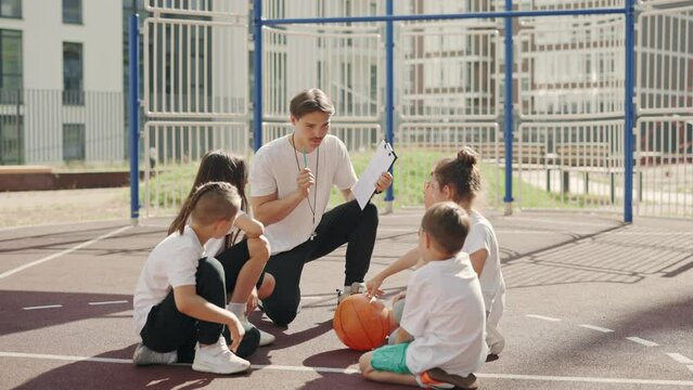 School coach explaining the rules of basketball game to children on basketball court. Children's basketball team in the school yard. Education and sport concept.