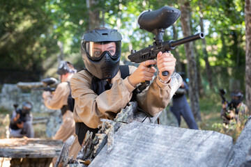 Fototapeta premium Girl in protective paintball outwear standing behind cover, aiming and shooting with paintball marker.