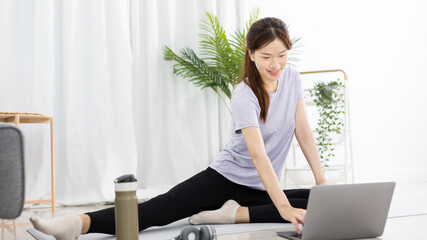 Beautiful Asian woman watching yoga teacher and light exercises in her living room at home, Watch live teaching or VDO on laptop, Dumbbells and a protein shake or bottle of water, Healthy exercise.
