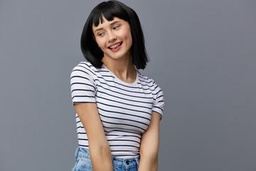 a sweet, pleasant, attractive, shy woman with black hair stands on a gray background in a white striped T-shirt and looks away