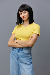  beautiful, attractive woman is standing in a yellow T-shirt and light jeans on a gray background with her arms crossed over her chest and smiling prettily at the camera. Vertical Studio Photography