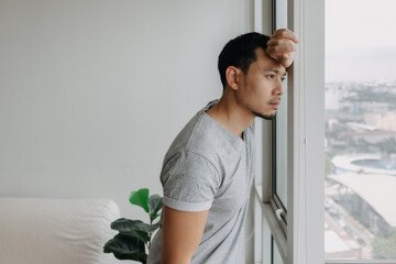 Thinking serious lonely asian man stand lean on the window in his apartment