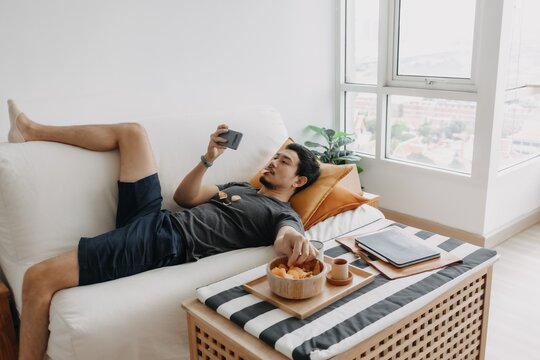 Asian Man Watch Online Tv And Eating Snack Instead Of Working From Home.