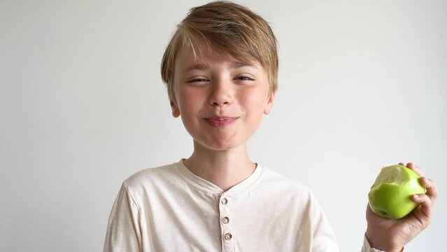 Laughing cute child boy bites green sour apple, chewing, smiling at camera over white background. Healthy lifestyle and eating habit for teens and children. Slow motion.