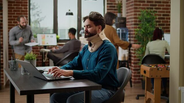 Hurt company employee with cervical collar after physical accident, working in pain at business office. Injured man wearing medical neck foam brace to cure back tension and fatigue.