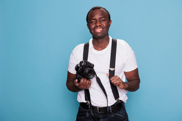 Smiling heartily professional photographer having DSLR camera while standing on blue background. Confident and handsome looking young adult person wearing trendy clothes while having photo device.