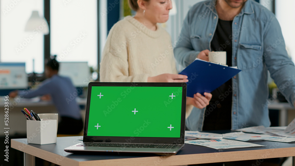 Poster laptop computer with green screen on desk in front of startup colleagues talking about sales charts 