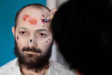 Artist using makeup and costume to create zombie look, sitting in studio. Man waiting to look...