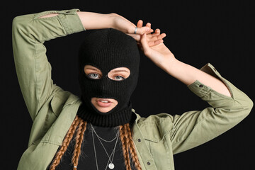 Portrait of young woman in balaclava with hands above her head on black background