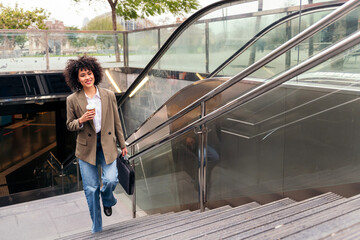 latin business woman smiles leaving the subway station with a briefcase and a coffee in her hand,...