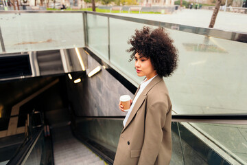 latin business woman entering the subway station on the escalator with a coffee in her hand,...
