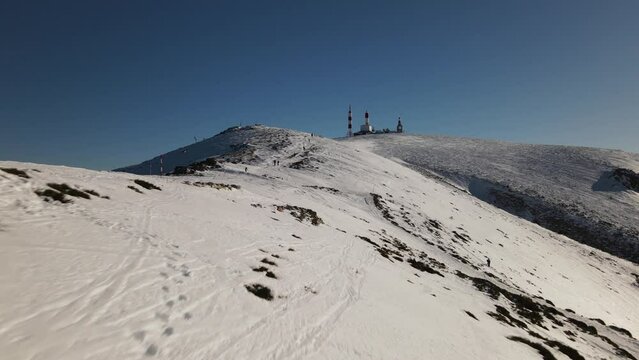 Push in aerial drone shot flying towards an antenna station as hikers passing by on the top of a snowy mountain.
