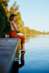 A middle-aged woman is relaxing on the dock and doing yoga breathing exercises. She put her feet in...