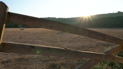 Uncovering a recollected field with round bales of wheat in the rising sun. Slow dolly vertical camera movement between the wooden bars of a gate. Cinematic shot with sun flares. - Powered by Adobe