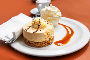 Serving of cheesecake on a white plate in focus. Small coffee cup out of focus in the background on orange color table. Tasty dessert.