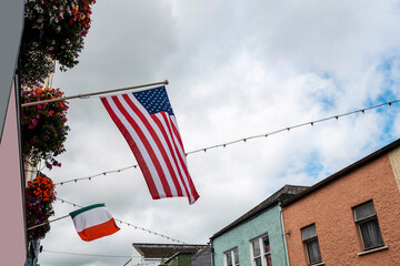 Flag of United States of America and Ireland on a building. Small colorful building in the background and cloudy sky. Relationship and history between two counties.