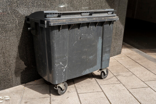 Black dirty trach wheelie bin in a street. Rubbish collection. Disposal of waste in a city.