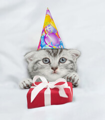 Cute kitten wearing birthday cap lying under blanket on a bed at home and holds gift box. Top down...