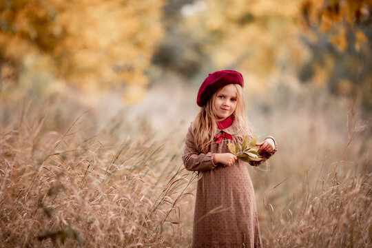 cute little blonde girl with long hair in autumn in the park smile and enjoys sunny day and good weather. preschooler in burgundy beret holds bouquet of yellow leaves. autumn walks and leisure