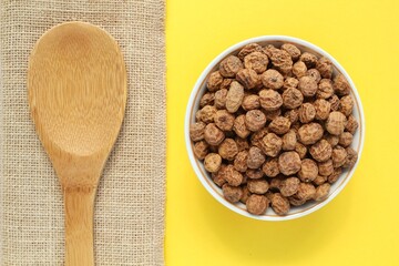 Chufa, tigernut, earth almond on white ceramic bowl and eco canvas bag near wooden spoon on yellow background. Healthy vegan food concept. Tiger nut for flour, oil, milk, traditional drink horchata