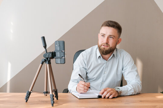 Cheerful bearded young man leading an online meeting using smartphone writing text information in notebook while sitting at table in an office in shirt. Positive guy talking on video link taking notes