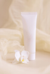 Obraz na płótnie Canvas White Cream tube near orchid flowers and tulle on light beige close up. Cosmetic packaging Mockup