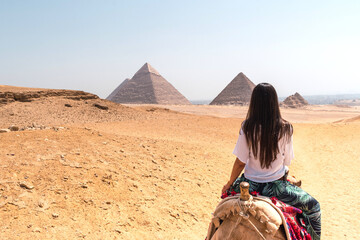 Woman with long dark hair mounted on a dromedary on her back looking at the pyramids of Egypt - obrazy, fototapety, plakaty