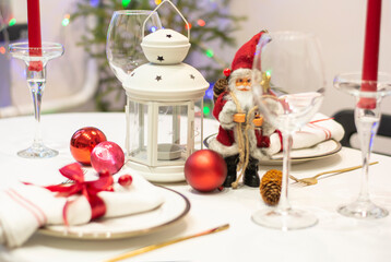 Stylish christmas decorated table plates with golden cutlery, napkin tied with red ribbon, red candle, santa claus figure, candlestick, wine glasses in classic style on christmas tree bokeh background