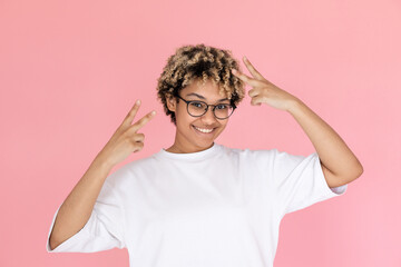 Portrait of happy mixed race woman making peace gesture. Young woman wearing eyeglasses and white T-shirt looking at camera and smiling against pink background. Success concept