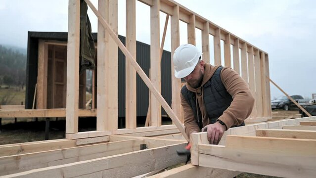 Man worker building wooden frame house on pile foundation. Carpenter hammering nail into wooden board, using hammer. Carpentry concept.
