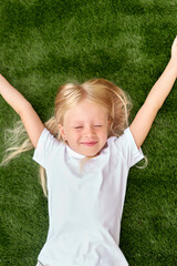 Pretty blonde girl lying and stretching on green grass. Mock up white t shirt. Top view.