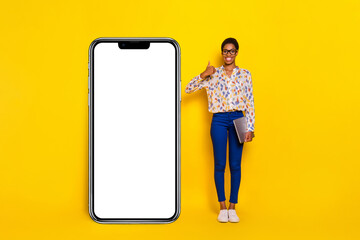 Full length portrait of positive person show thumb up empty space huge phone isolated on yellow color background