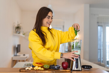 Young woman making smoothie with fruits. Beutiful girl standing in the kitchen and preparing smoothie with fruit and vegetables