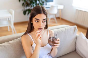 Concept of suffering from terrible pain. Beautiful, sad, upset, unhappy, troubled, weak woman with black hair, wearing casual clothes is sitting on a sofa and taking a pill