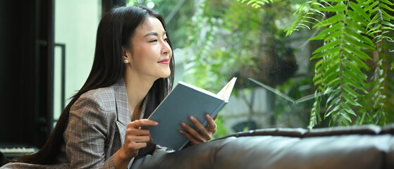 Pleasant millennial business woman resting on couch in bright comfortable office and reading book