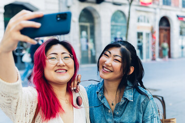 Two pretty young Asian women with a big smile taking a selfie with their smartphone.