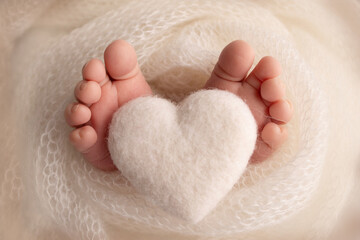 Knitted white heart in the legs of a baby. Soft feet of a new born in a white wool blanket. Close-up of toes, heels and feet of a newborn. Macro photography the tiny foot of a newborn baby.