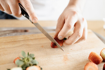 A man wearing a white t-shirt chopping up strawberries in a kitchen on a wooden board with a knife