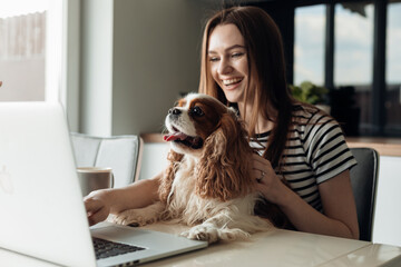 Delightful smiling young woman sitting in kitchen with dog coker spaniel, use laptop. Video call to...