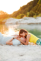 A young couple in love are resting on the shore of a lake, lying on the sand in summer sunny weather at sunset.