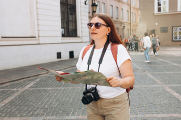 Attractive young female tourist with camera is exploring new city. Redhead girl holding a paper map in a sunny day. Urban lifestyle concept. Traveler, old town